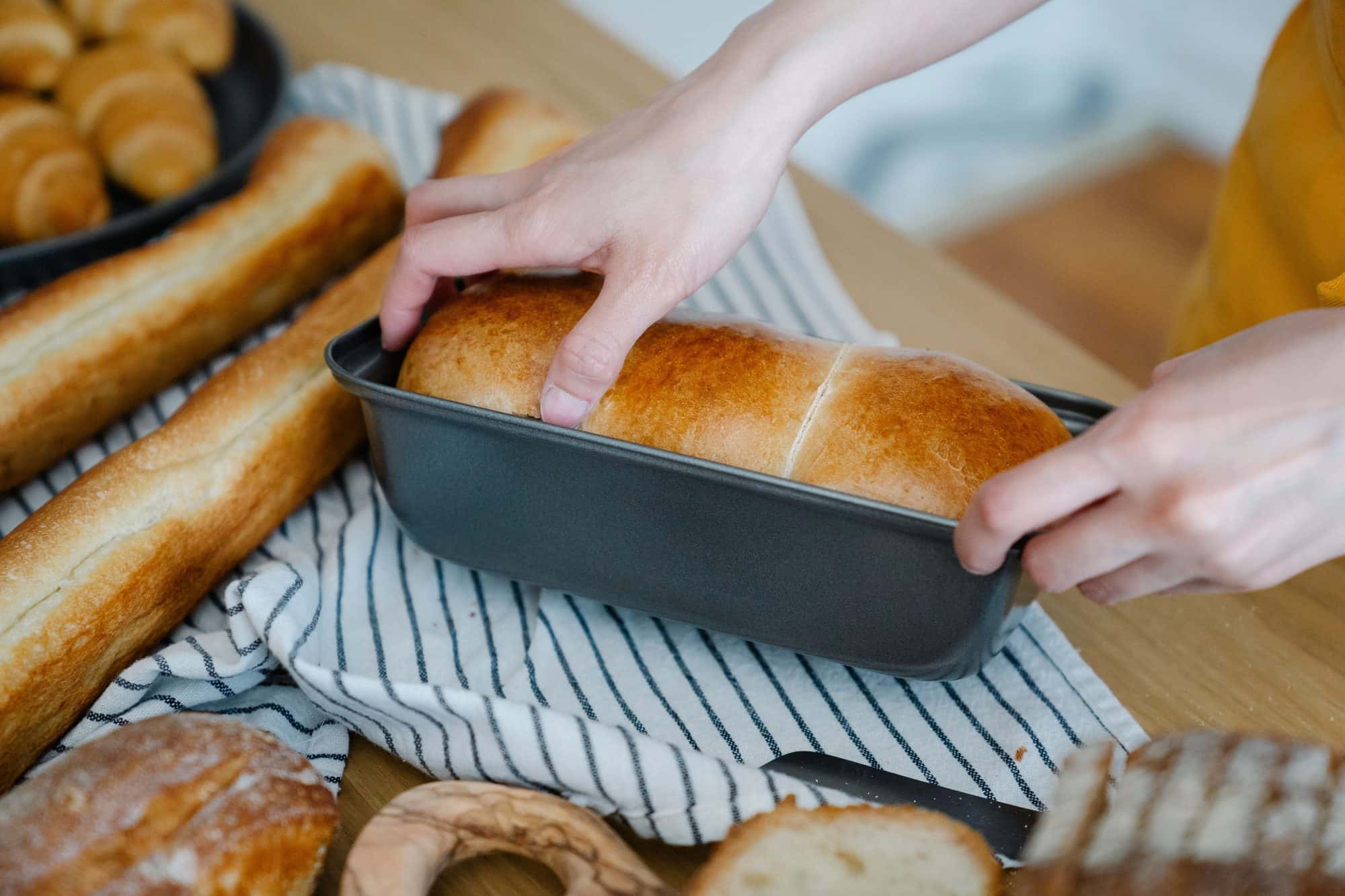 Handen halen een vers brood uit de vorm, naast stokbroden en brood op een houten tafel